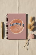 Manifest It dusty pink spiral notebook upright on cream linen with pampas grass and rose quartz