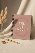 SHE IS ENOUGH dusty pink spiral notebook upright on cream linen with gold pen and pampas grass